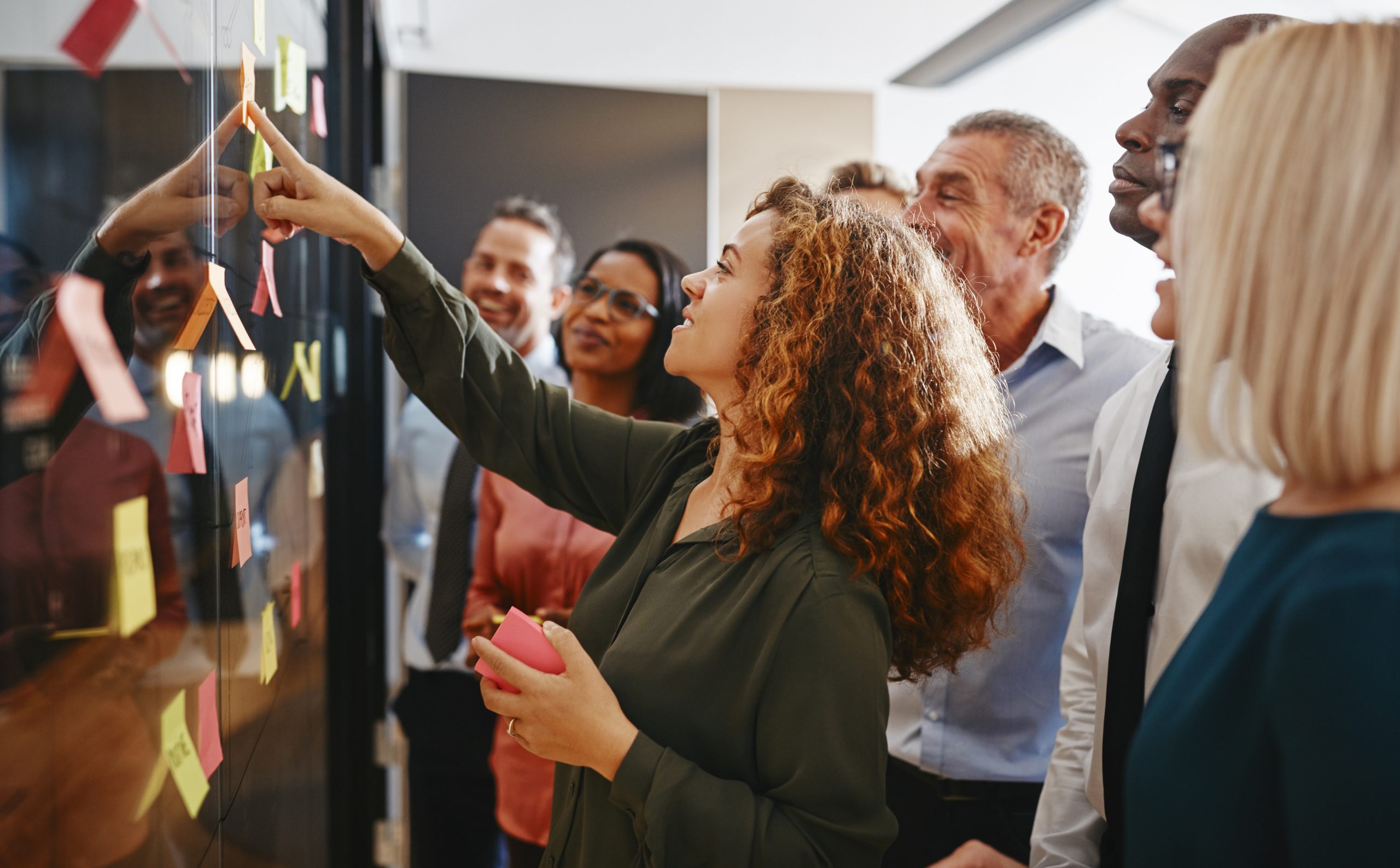 A group of people in the office, discussing and pointing at sticky notes on a glass wall.