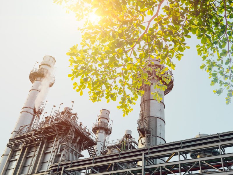 Factory on a sunny day with foliage in the foreground. Illustrates the intersection between business operations and the environment. A way of thinking about double materiality.